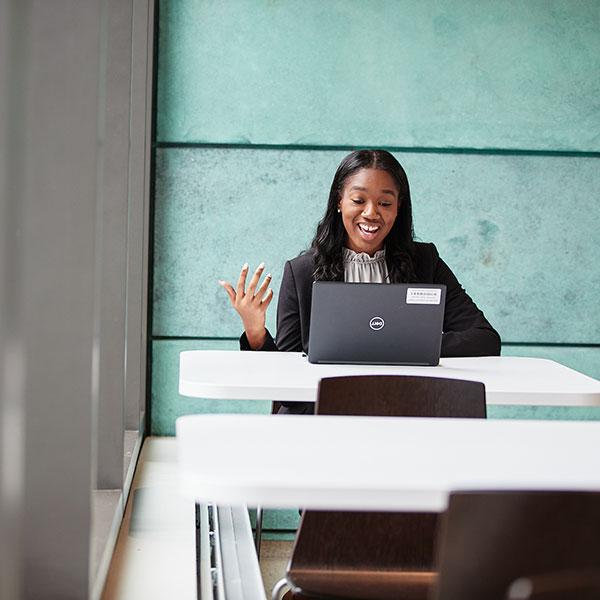 A student sits at a table and video conferences from her laptop. She’s smiling and speaking to the screen.